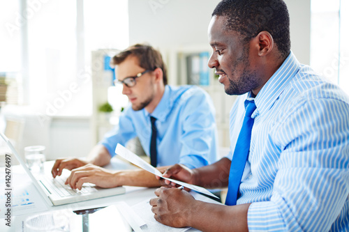 Middle-aged jurist in eyeglasses drawing up agreement on laptop while his Afro-American boss with document in hands keeping eye on him