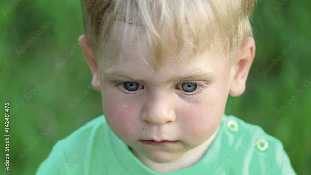 Closeup portrait of pensive blond caucasian boy over green grass background. Little child of 1 year and 5 month age thinks about something ignoring everything around. Video shot from above. Closeup portrait of pensive blond caucasian boy over green grass background. Little child of 1 year and 5 month age thinks about something ignoring everything around. Video shot from above.