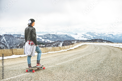 Stylish red-haired girl in a cap and denim overalls standing on a bump at the highway in the mountains with a longboard