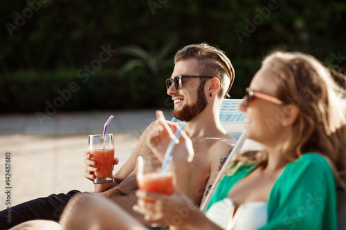 Friends smiling, drinking cocktails, lying on chaises near swimming pool.