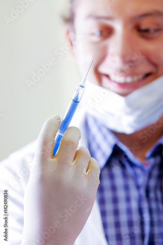 Young doctor with long dread locks posing for camera holding syringe  wearing facial mask covering mouth  clinic in background  medical concept
