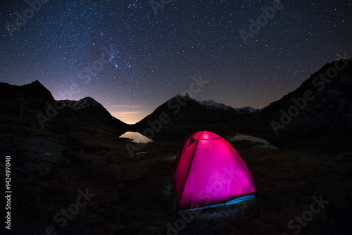 Camping under starry sky and Milky Way arc at high altitude on the italian french Alps. Glowing tent in the foreground. Adventure into the wild.