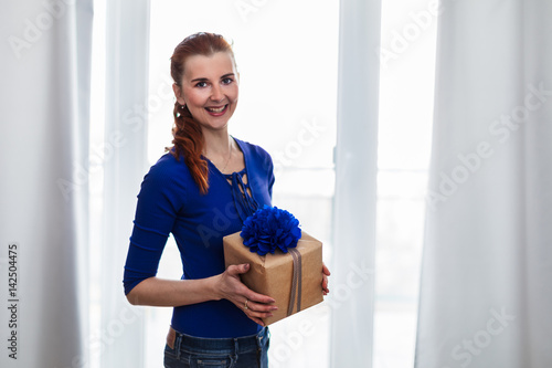 Smiling woman standing in front of the window holding a blue gift box.