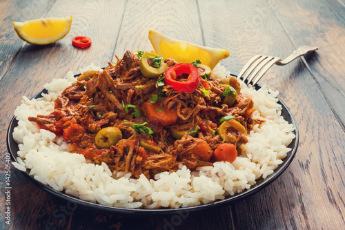 Slow Cooker Ropa Vieja, Cuban famously delicious shredded beef stew with pepper and olives on a black plate with a boiled rice and fresh lemon on the wooden table.