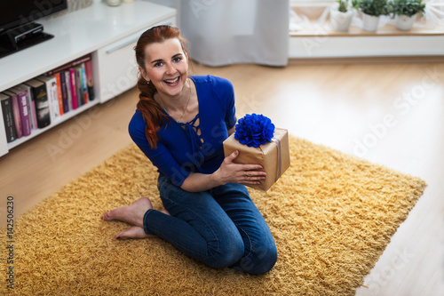 Smiling woman sitting on the floor holding a blue gift box. Upper view.