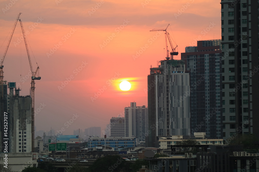 Fototapeta premium Sun setting on pastel orange sky amongst working cranes of the construction site, Bangkok, Thailand 