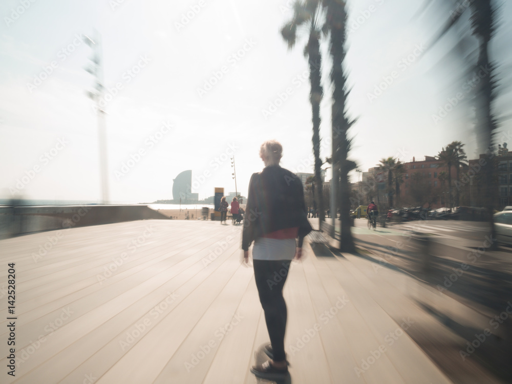 Young blonde modern woman with leather jacket riding her skateboard at the boardwalk of Barcelona motion blur