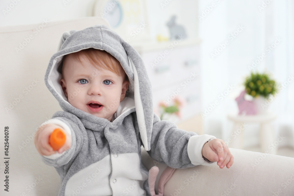 Cute little baby in bunny costume with carrot in hand sitting on armchair at home Cute little baby in bunny costume with carrot in hand sitting on armchair at home