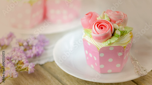 Vanilla cup cake and spoon on a plate with cup cake in background and flower as decoration.