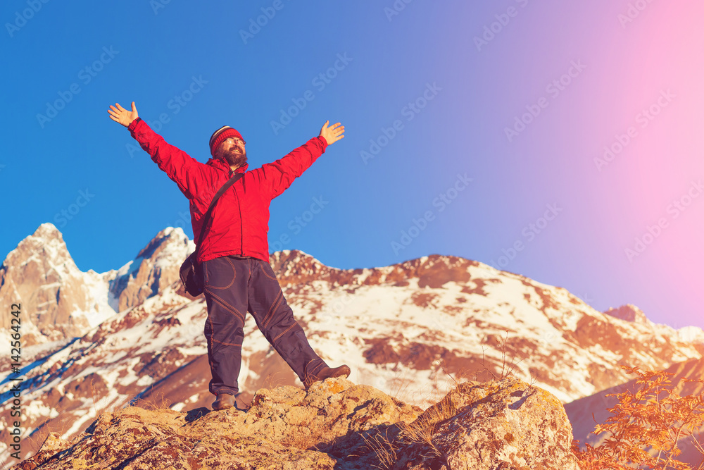 Obraz premium Bearded man in a red jacket standing with hands up in the mountains in the sunlight