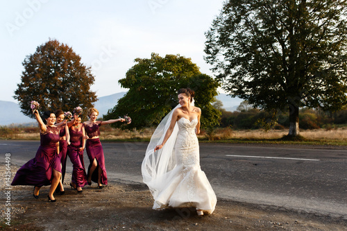 Bride and funny bridesmaids run along the road in sunny evening