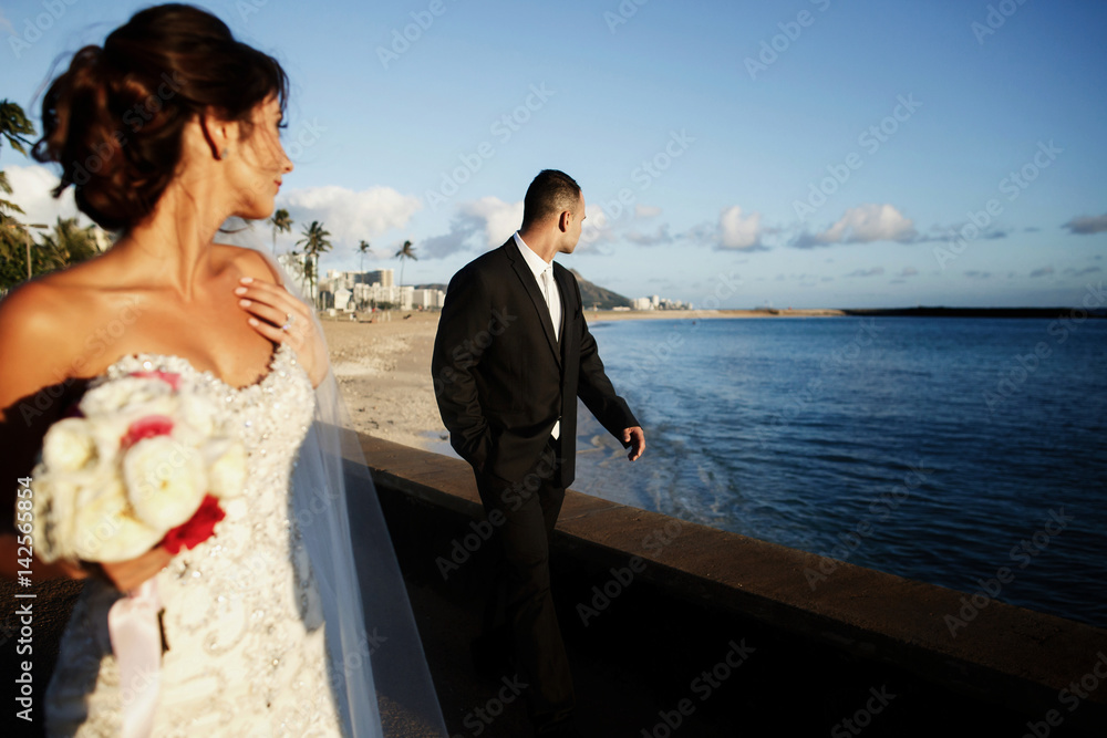 Bride looks over her shoulder at groom standing on ocean shore Bride looks over her shoulder at groom standing on ocean shore