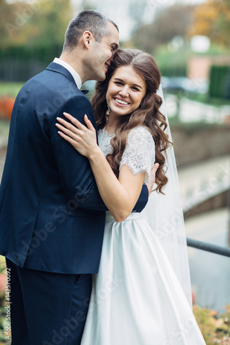 Pretty bride smiles while groom kisses her forehead hugging her © myronovychoksana