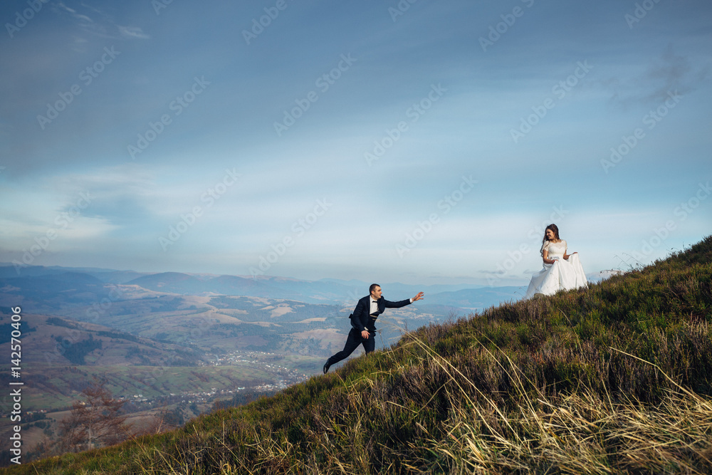 Groom reaches his hand out to the bride walking behind her on the hill