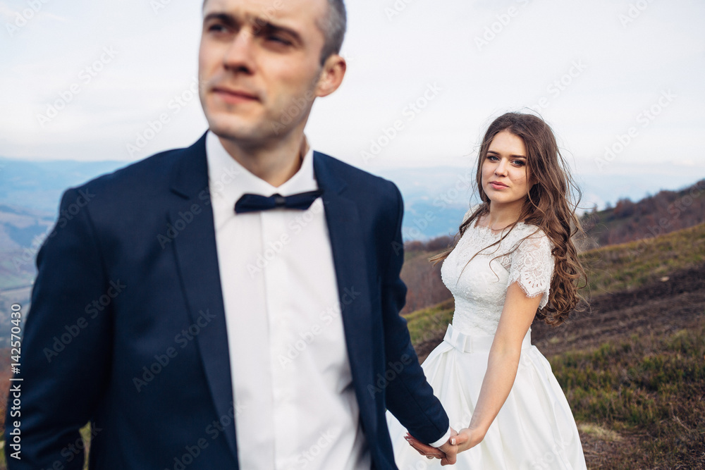 Curls hang on bride's shoulders while she stands with confident groom on high hill Curls hang on bride's shoulders while she stands with confident groom on high hill