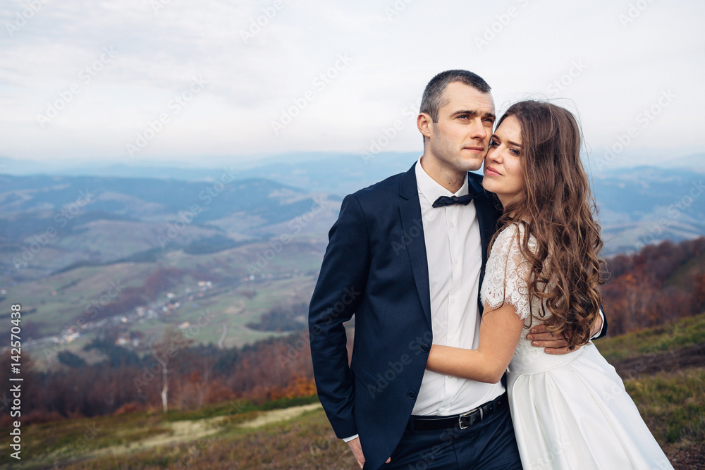 Tall groom in black suit holds bride tender standing under deep autumn sky