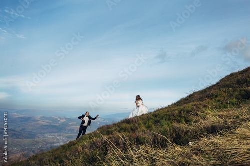 Groom reaches his hand out to the bride walking behind her on the hill © myronovychoksana