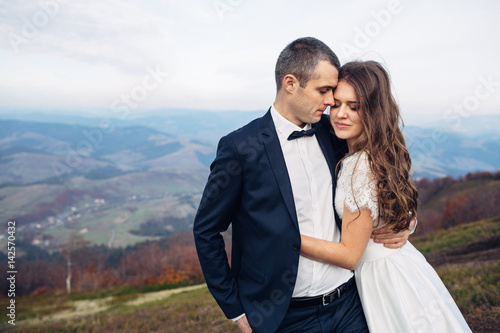 Tall groom in black suit holds bride tender standing under deep autumn sky