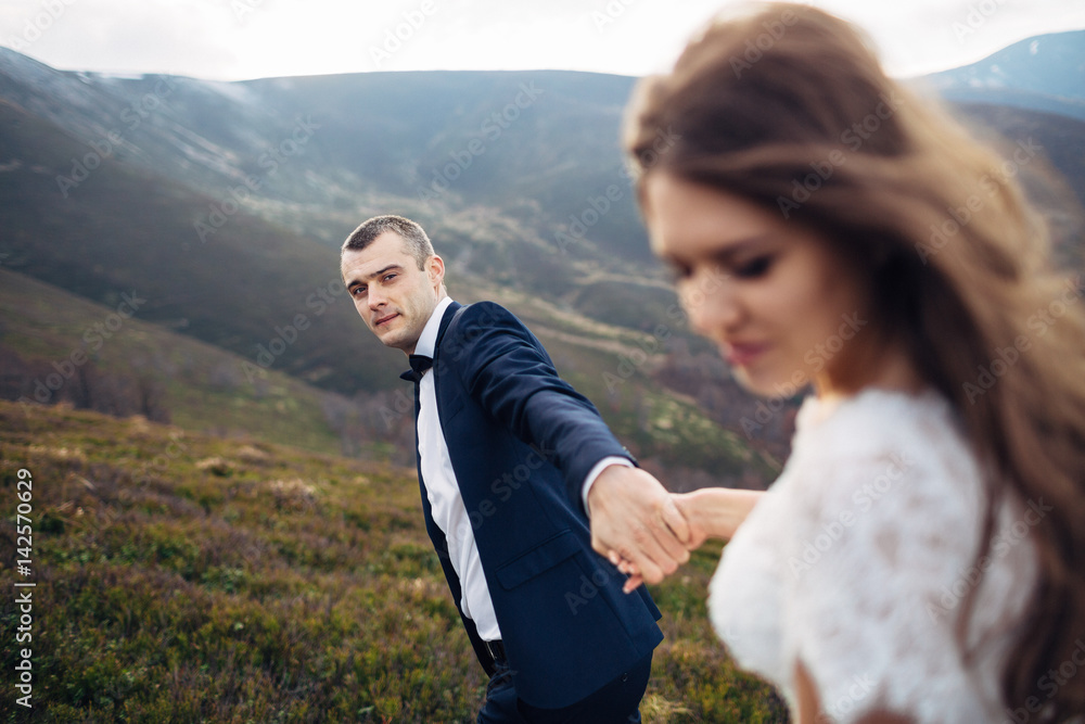 Groom looks at bride holding her hand on windy mountain hill
