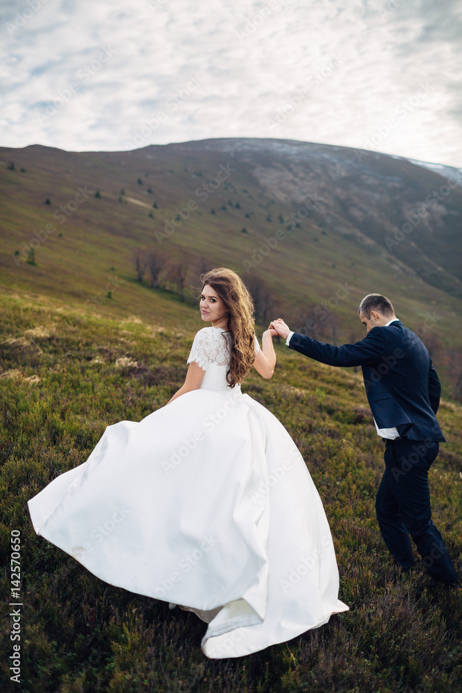 Wind blows bride's magnificent dress while she climbs hill together with groom