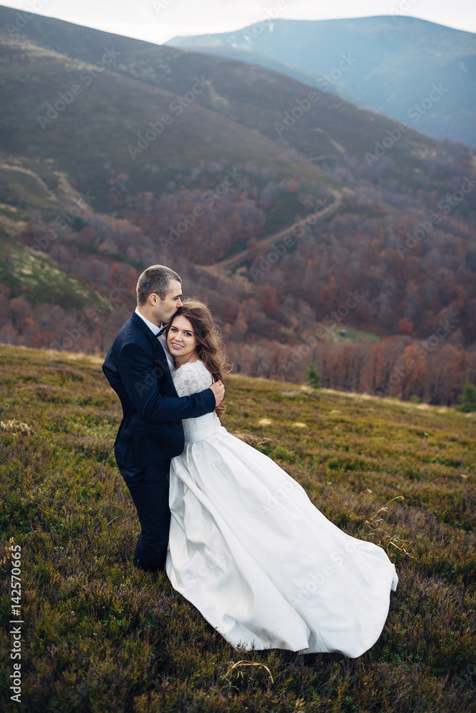 Groom kisses bride's head hugging her on windy hill