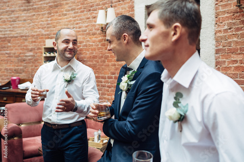 Groom and groomsmen chat drinking whisky in old brick hall photo