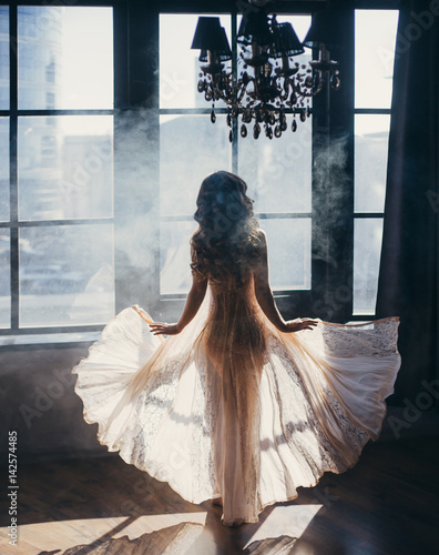 The girl stands with her back to the camera on the background of a huge window. Transparent dress flies to the sides. photo