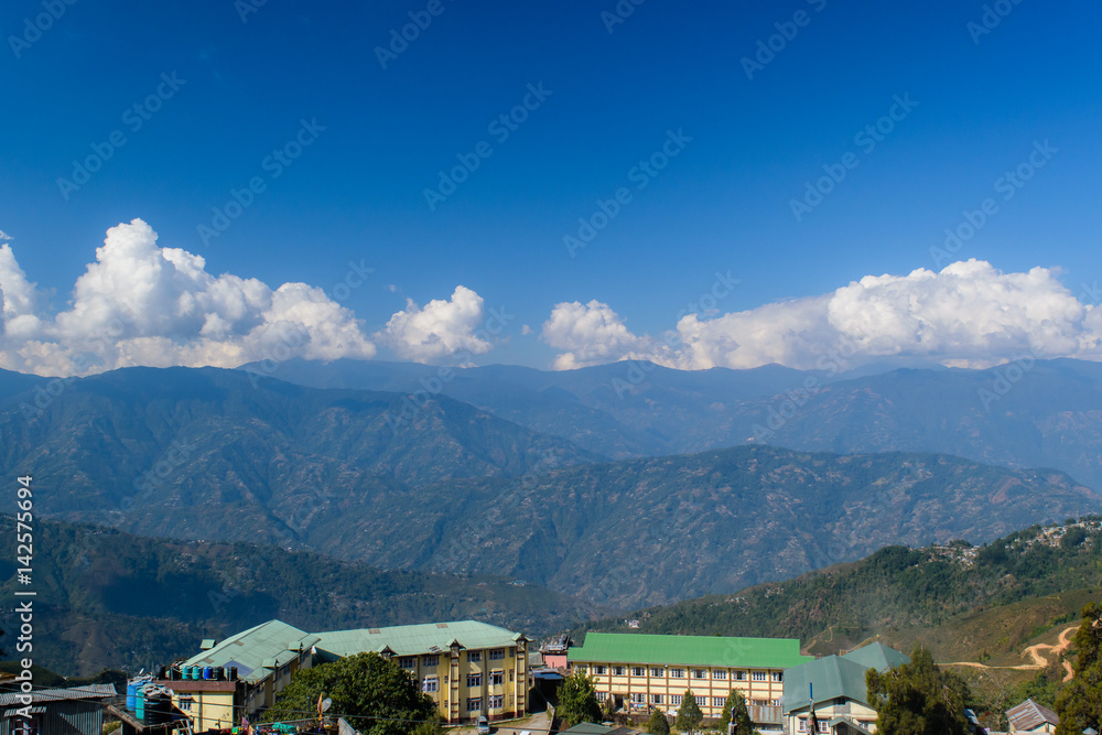 A hilltop view of house' roof with mountains and blue sky with clouds from Darjeeling Himalayan railway station on a misty morning.