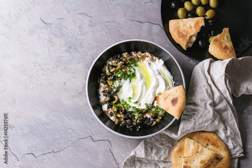 labneh middle eastern lebanese cream cheese dip with olive oil, salt, herbs, olives tapenade served in black bowl with traditional pita bread over gray texture metal background. Top view with space