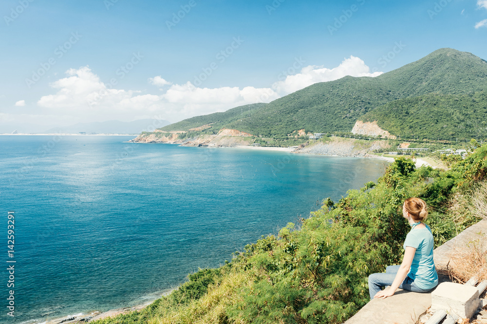 Woman sitting on the brink of break. Calm water in sea bay with rock, bush and green forest and clouds in background