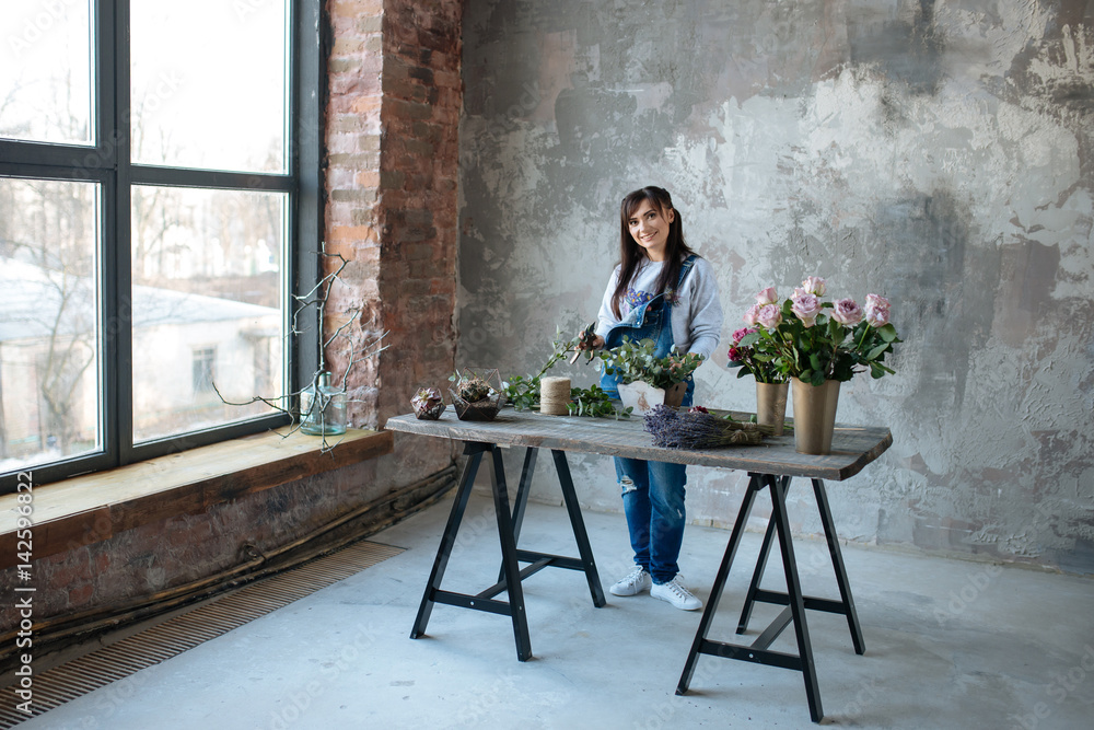 female in gray blouse and jeans make a bouquet over gray background, putting roses in vase, flowers and vase on wood table, workplace female in gray blouse and jeans make a bouquet over gray background, putting roses in vase, flowers and vase on wood table, workplace