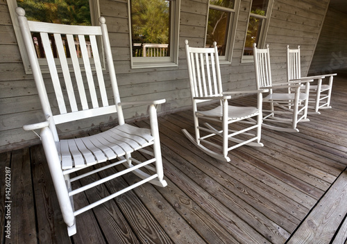 Four white rocking chairs on wooden porch.