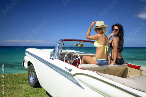 Two beautiful young women brunette and blond in swimsuits sit into retro car cabriolet near caribbean coastline