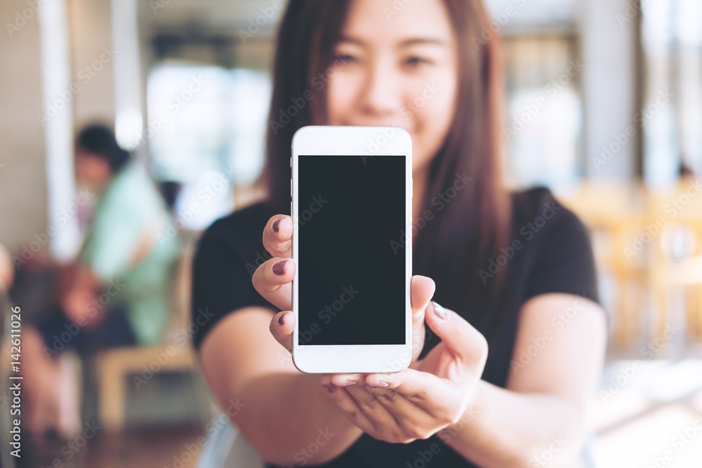 Mockup image of a beautiful woman holding and showing white mobile phone with blank black screen and smiley face in modern cafe