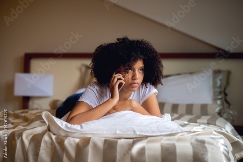 Cuadro en lienzo Black girl lying on bed making phone call, looking away.