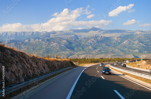 Several cars on a highway in Croatia against a rocky mountain range on a warm summer day. Blue sky with fluffy white clouds on the back