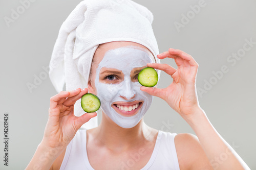 Young woman with facial mask holding slices of cucumber on gray background.