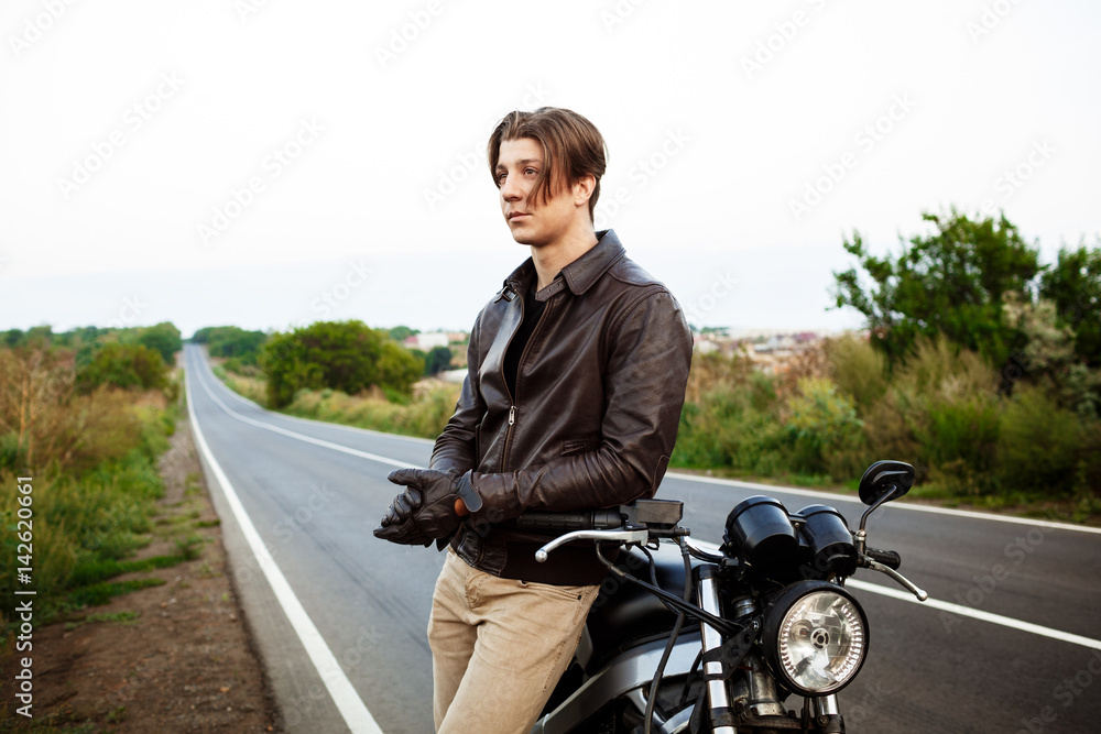 Young handsome man posing near his motorbike at countryside road.