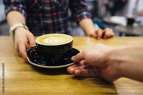 Cropped image of two people's hands at a table with cup of cappuccino. Barista serving coffee to female customer in cafe. Close up of human hands touching with cup
