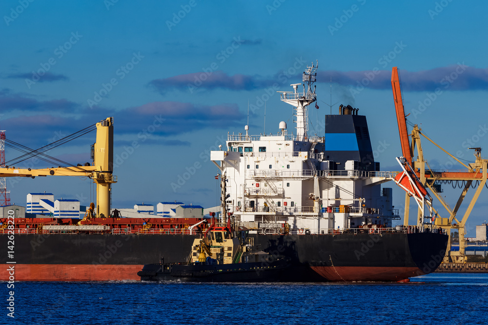 Fototapeta premium Black cargo ship mooring at the port with tug ship support