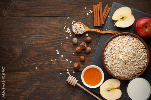 Raw oat flakes in bowl with various ingredients for breakfast on the dark rustic wooden background.