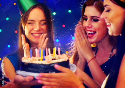 Happy friends birthday party with candle celebration cakes. Girl joyfully clapping. Small group people looking at burning candles in night spot. Women have fun hen-party on dark background.