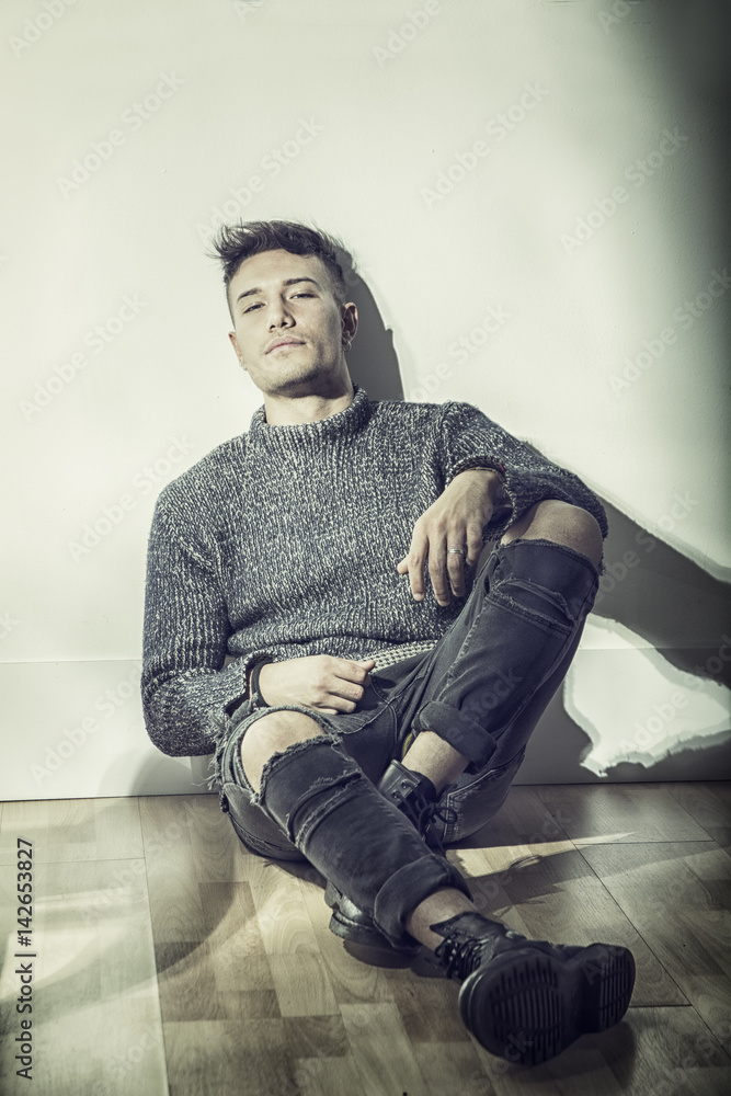 Fototapeta premium Handsome white asian young man wearing grey sweater and ripped jeans, sitting on light background against wall in studio shot