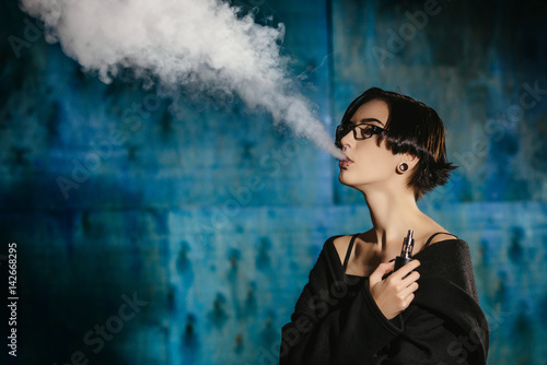 A girl with glasses vaping and releases a cloud of vapor. Model in a black vaper smoke vaporizer on a turquoise background