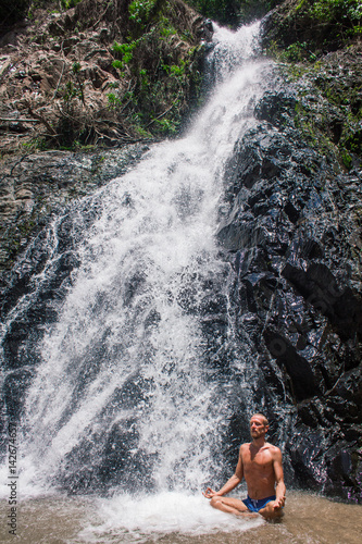 Man practicing yoga among waterfall. Young male sitting in lotus pose on rock with waterfall streams near in tropical forest.
