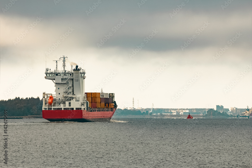 Fototapeta premium Red cargo container ship entering the port of Riga in cloudy day