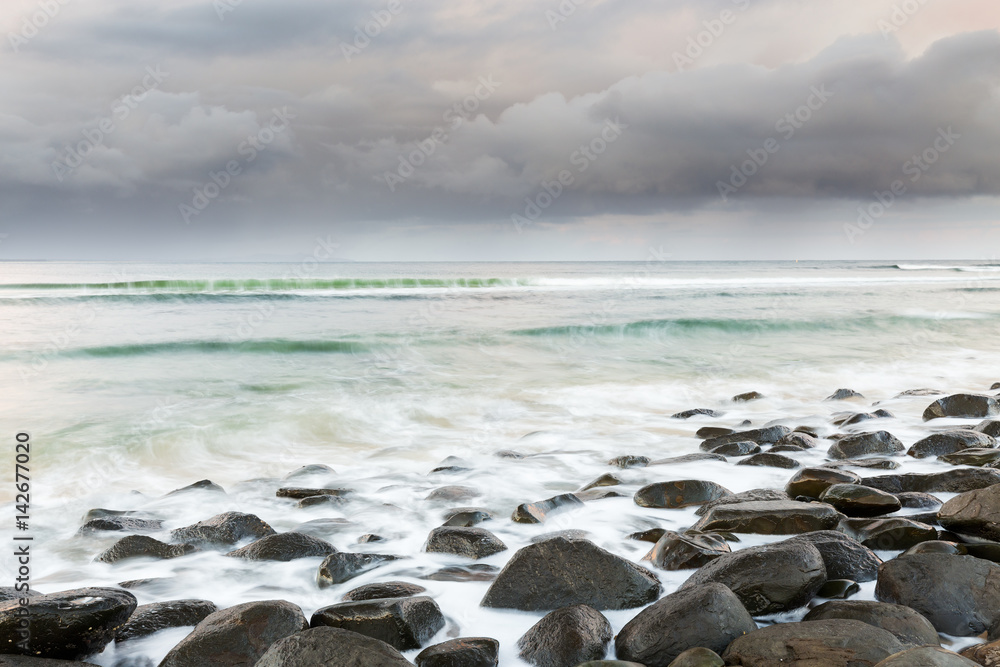 A storm passes on the horizon as waves wash over a boulder strewn beach in this seascape scene.