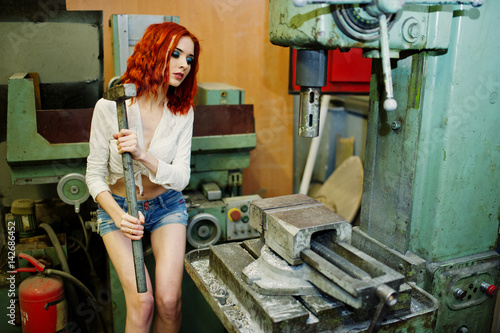 Red haired girl wear on short denim shorts and white blouse with hammer at hands posed at industrial machine at the factory. photo