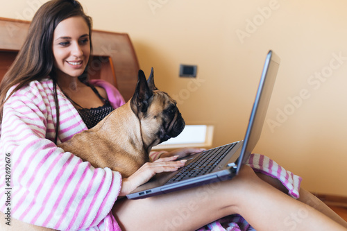 Attractive young woman sitting on bed with her french bulldog pet and doing something on laptop.