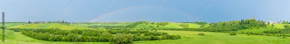 Obraz premium Wide panorama of hilly landscape with full rainbow in the sky, villages and church in background. 